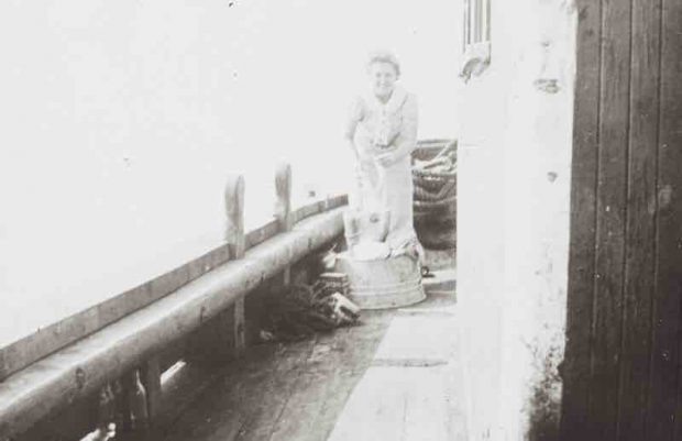 A black and white photograph. A young smiling woman, hair tied back, is standing on the deck of a ship. She appears to be washing dishes in steel pails. There are a few bundles of rigging all around her.