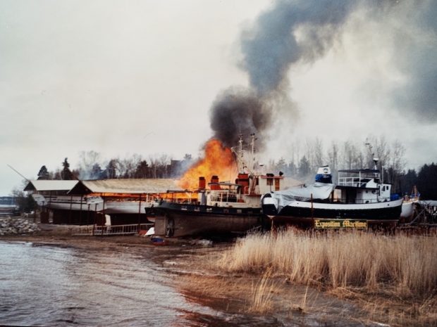 View of a riverbank with four ships. In the middle, flames and grey smoke rise out of a wooden ship. In the foreground a sign in the brown grass reads: “Exposition maritime”.