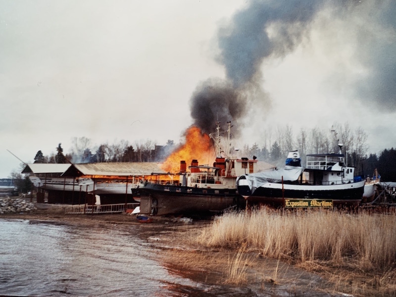 Vue d’une berge et de quatre bateaux. Au centre, des flammes et de la fumée grise s’échappe d’un bateau de bois. En avant plan, des herbes brunes et un panneau indiquant « Exposition maritime ». 