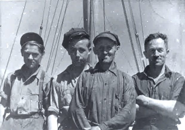 A black and white photo of four men in work clothes. On the left, two teenagers, in the middle, an older man, and on the right a man in his prime. Behind them we can see two masts and some rigging.