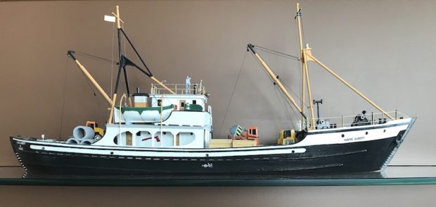 Model of the Havre-Aubert schooner. A small wooden model of a ship. The lower portion is black with the upper portion painted white. Small lifeboats and miniature construction materials complete this model ship. The name of the schooner is written on its bow.