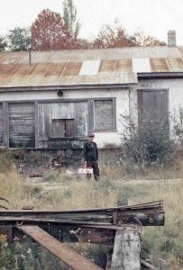 An elderly man is standing in the centre of the frame. He is carrying a bushel of apples. There are tall grass and a structure made of rusty metal and wood in the foreground. A boarded-up building is visible in the background.