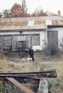 Un homme âgé se tient au centre de l’image. Il a dans sa main un panier rempli de pommes. Devant lui, des herbes hautes et une structure en bois et en métal rouillé. Derrière lui se dresse un bâtiment placardé. 