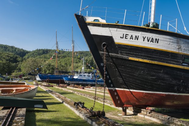 A photograph of the site of the Musée maritime de Charlevoix. In the foreground we see part of the Jean-Yvan schooner’s hull, painted white, black, yellow and red. Rails form straight line leading to two more boats adorned with flags. To the left, a couple of rowboats are resting on the ground.