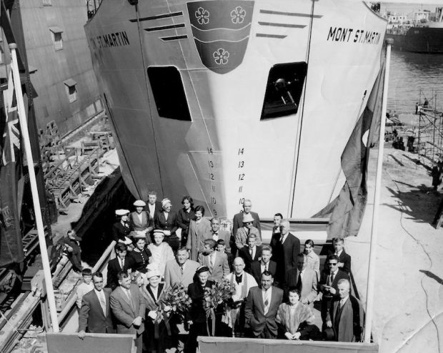 Black and white photograph. In the foreground, a group of people in Sunday clothes on a small stage. In front of them, a priest is standing next to two women holding bouquets of flowers. Behind them, towering over the group, is the huge white hull of a ship. The name of the ship is painted on both sides of the hull: Mont St-Martin.