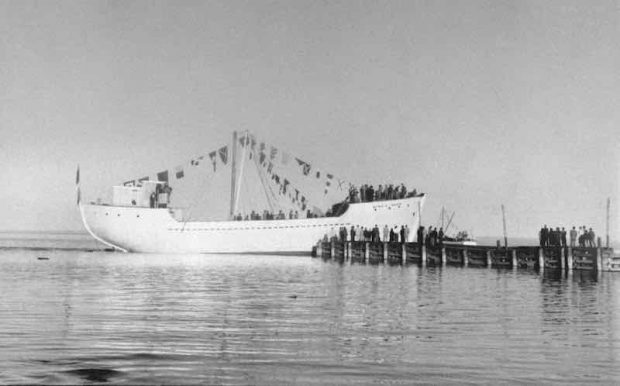 A black and white photograph of a schooner on the St. Lawrence River. There is a large body of water in the foreground. To the right, a dock stretches out into the water. Many people are standing on the dock, observing the majestic white ship adorned with many maritime flags. There are more people aboard the schooner.