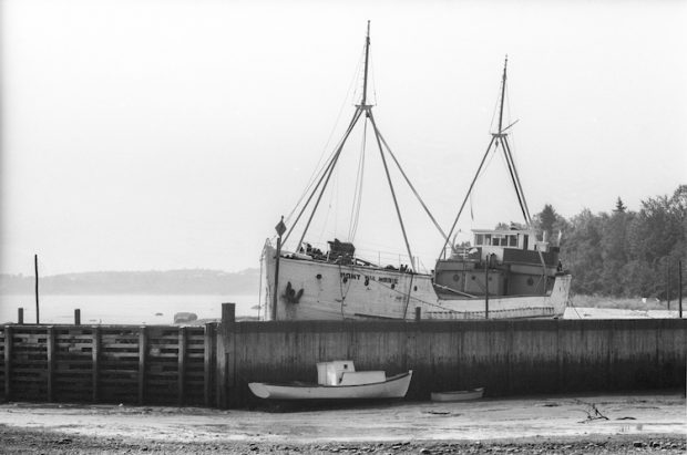 Black and white photograph. In the centre of the picture stands the Mont Ste-Marie schooner at dock. The ship is white, with its name painted black. It has two masts. The dock is made of wood. There are two small rowboats in the water in front of it.