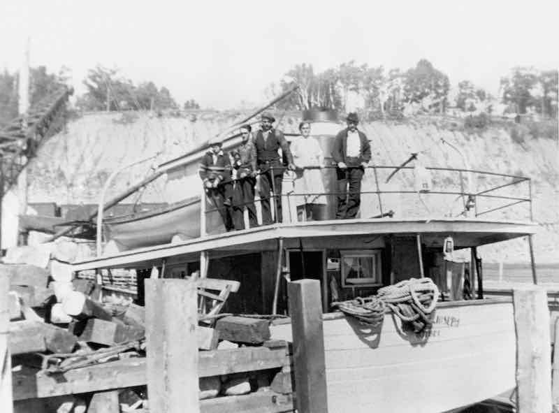 A black and white photograph with, to the left, a dock with lumber strewn about, and to the right, the Mont St-Joseph schooner. The name of the ship is partially hidden by the rigging. Four people are posing on the ship: three young men and their parents.