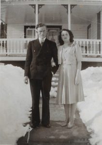 A young couple posing for the camera. On the left, the man is dressed in a three-piece black suit and is wearing a tie. On the right, the young dark-haired woman is wearing a light-coloured dress with a short jacket. Behind them we see a house and a lot of snow.