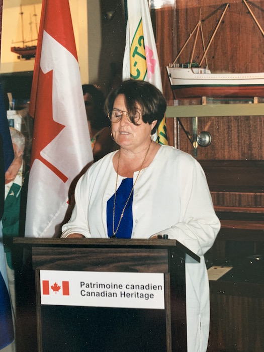A woman is standing behind a lectern featuring the logo of Heritage Canada. She is giving a speech. Behind her there are two flags and a scale model of a ship.