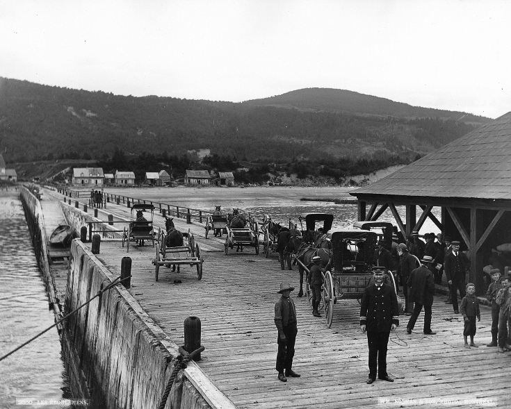 View of the dock at Les Éboulements (Saint-Joseph-de-la-Rive), circa 1890. This photo was taken from a ship moored at the end of the dock, the shore, along with a few houses are visible on the horizon. The wooden dock. In the foreground, a man stands in uniform. Next to him are a few children and other men. Some horse-drawn carriage in the background.