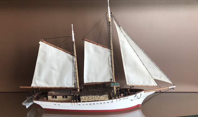 Model of the Rose-Alba sail-powered schooner. The hold of the ship is white, with a small red stripe at the bottom. The ship is fitted with miniature ropes and a small lifeboat at its stern. The model sports four white sails.