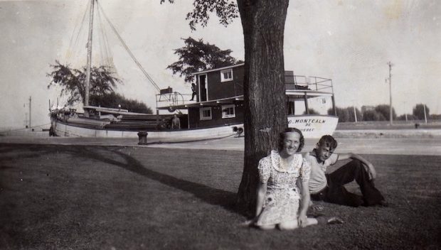 A black and white photograph. In the foreground, a young man and woman are sitting on the grass near a tree. Behind them there is a wooden ship, on which three people are barely discernible. The boat seems to float on a narrow canal.