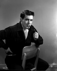 A black and white portrait of a young man He is sitting backwards on a wooden chair, with the backrest against his belly. The young man is wearing black, with a white scarf knotted around his neck and a pipe in his mouth.