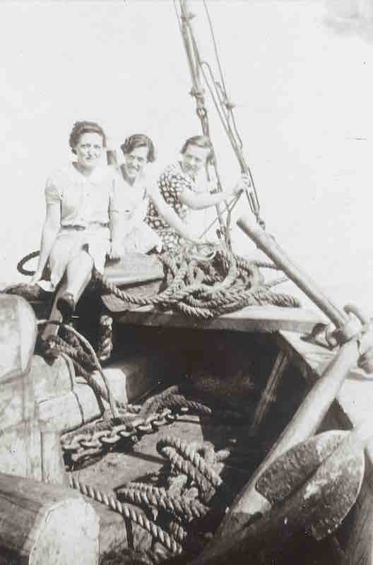 A black and white photograph of three young women holding on to a ship’s bow. In the foreground are some logs, rigging and an anchor.