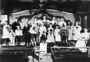 Christmas concert with children on stage at school holding garlands and small tree in front.