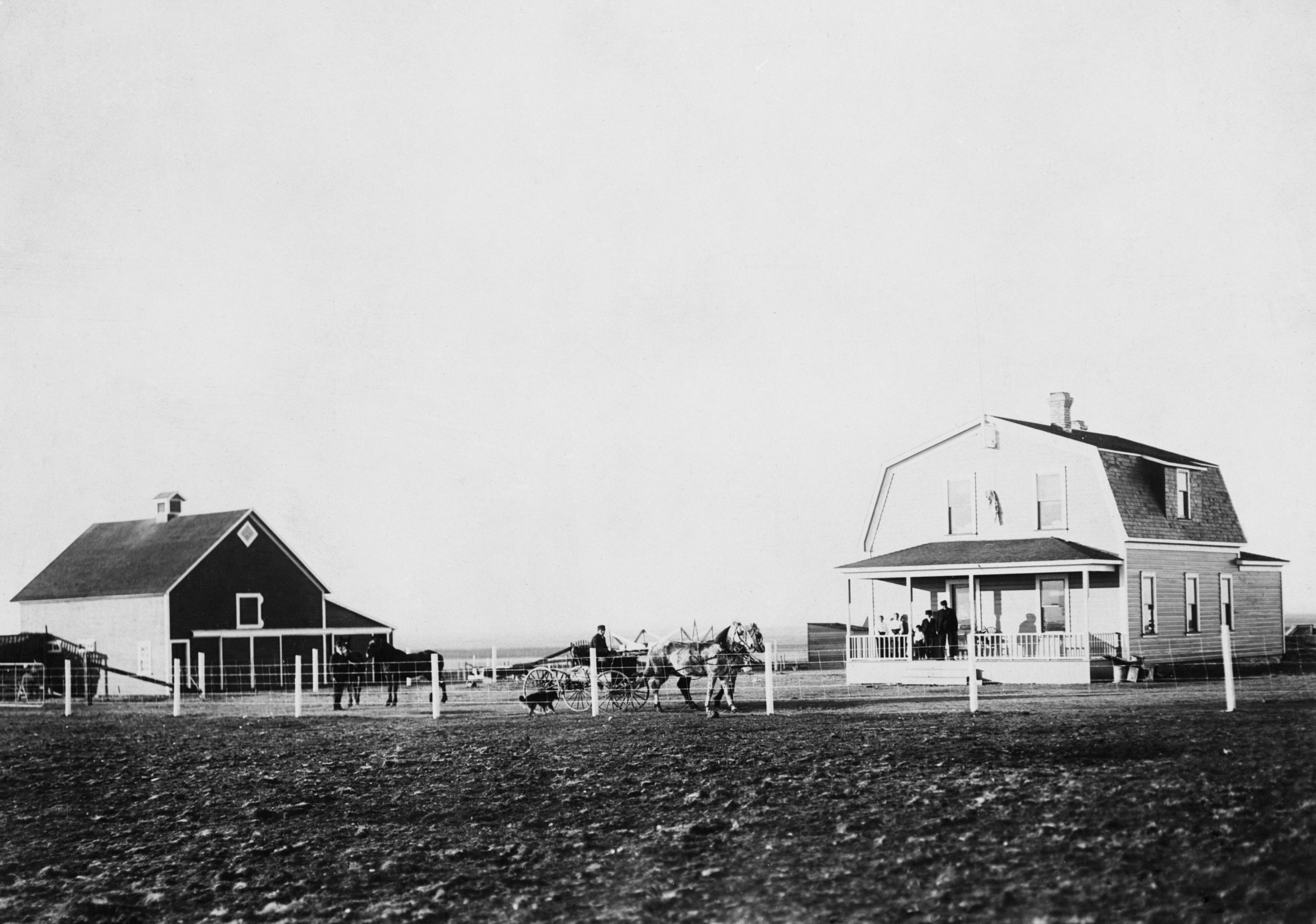 Large hip roof house with porch and dormer window. Fenced yard with barn, horses and buggy and a dog. Several people on porch, with horses and a man in the buggy.