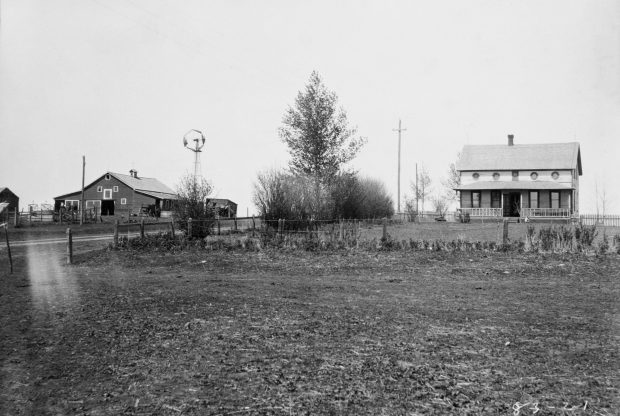 Farmyard with large two story house with porch, barn and outbuildings, windmill on treed, mowed and fenced yard.