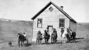 Cinq chevaux alignés devant une école, deux enfants sur chaque cheval. Un cheval et un chariot avec trois enfants et un chien.