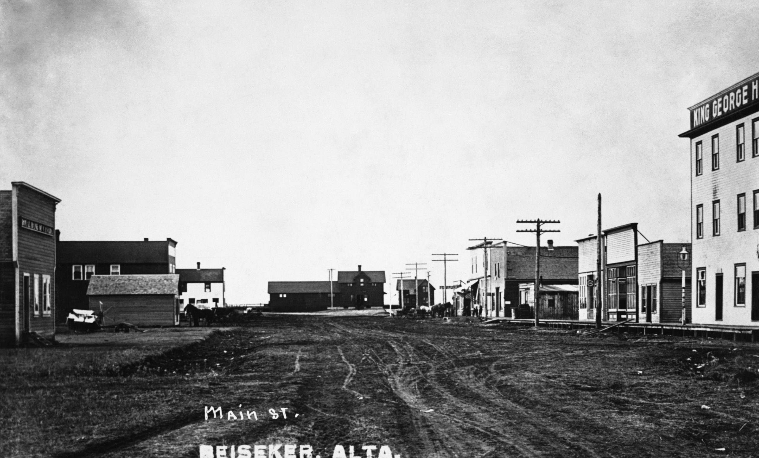 Main Street Beiseker. Wide dirt road with sundry businesses on each side, horses and buggies hitched outside. Right side includes the King George hotel and telephone poles. The street leads to the train station.