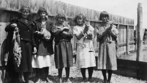 Five schoolgirls, with bobbed hair and mid length dresses, along a fence. Four are holding coyote pups, one is holding a string of snared gophers.