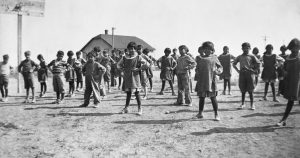 Groupe d’enfants autochtones dans la cour, les mains sur les hanches, avec bâtiment en arrière-plan.