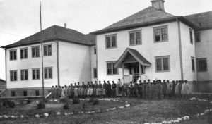 Large two story residential school, children and teachers lined up in front, large yard with stones in a circle and flagpole.