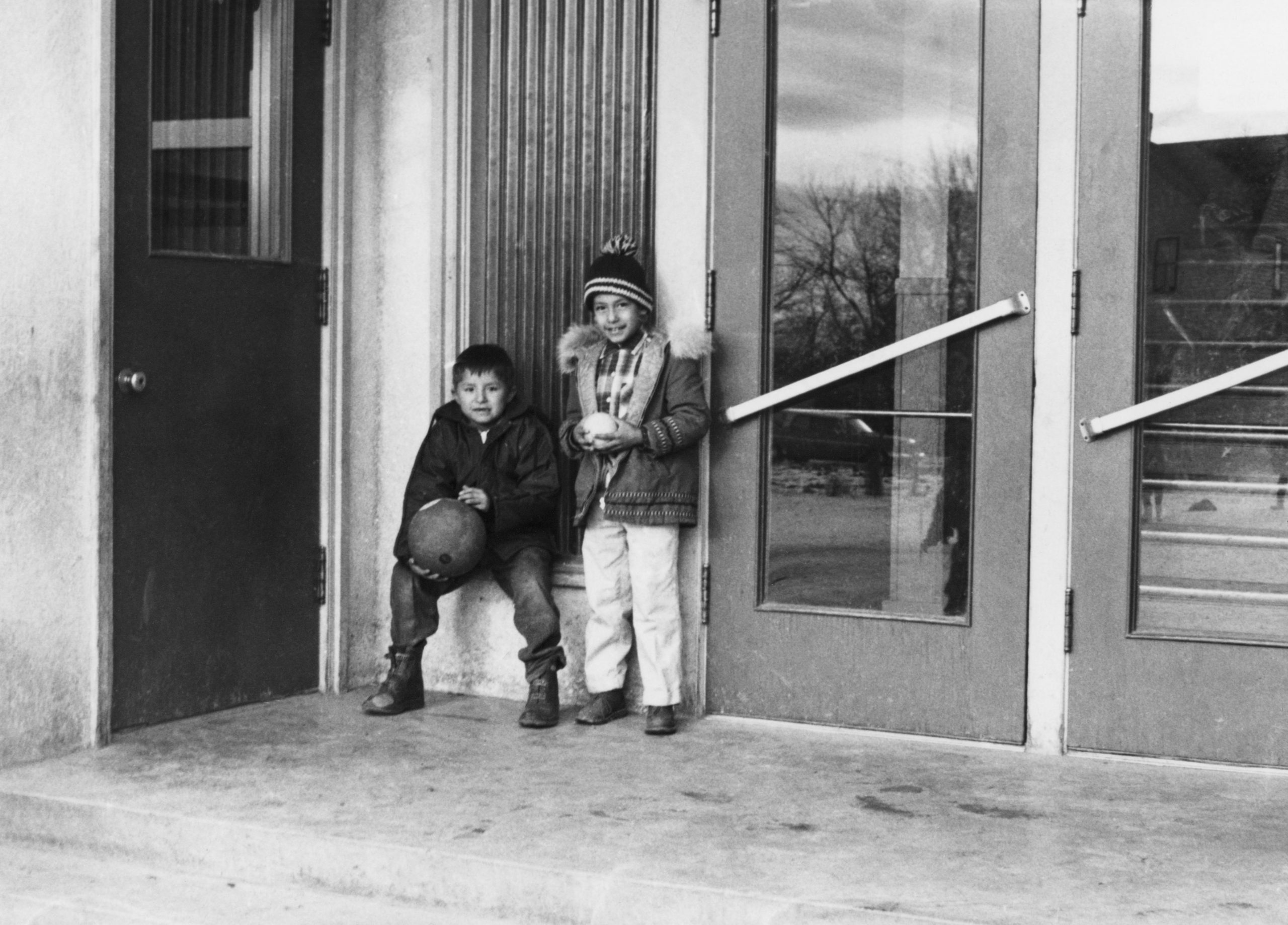 Two boys holding balls, dressed in winter clothes, boots, coats, toque, no gloves, standing outside front doors.