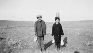 Two schoolboys standing by a fencepost in a field, holding syrup pails.