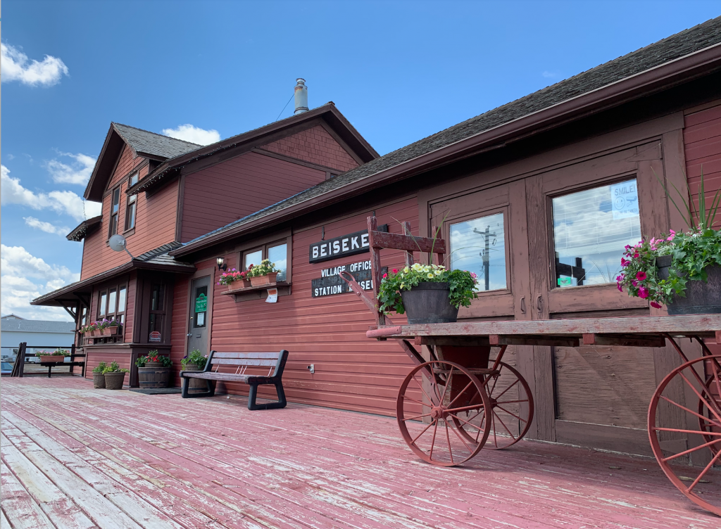 Entrance to the Beiseker Museum. Rust-colored two-story building with bench, flowerpots and steel wheeled cart with flowers.