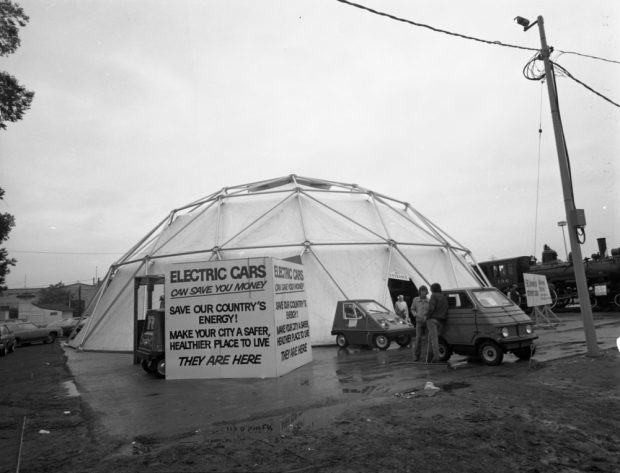 Black and white photograph of a geodesic dome structure, with several electric cars parked out front and a train visible in the background. Signage reads “ELECTRIC CARS / Can save you money / SAVE OUR COUNTRY’S ENERGY!”