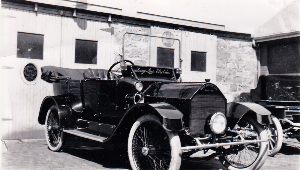 Black and white photograph of an automobile with a single headlight parked at a garage. Writing on the vehicle’s windshield reads “Storage-Gas-Electric”