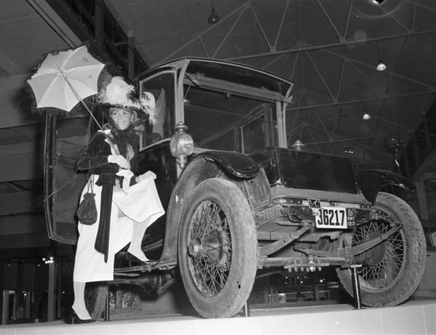 Black and white photograph of a model in early 20th-century dress posed in the door of a vintage electric car.