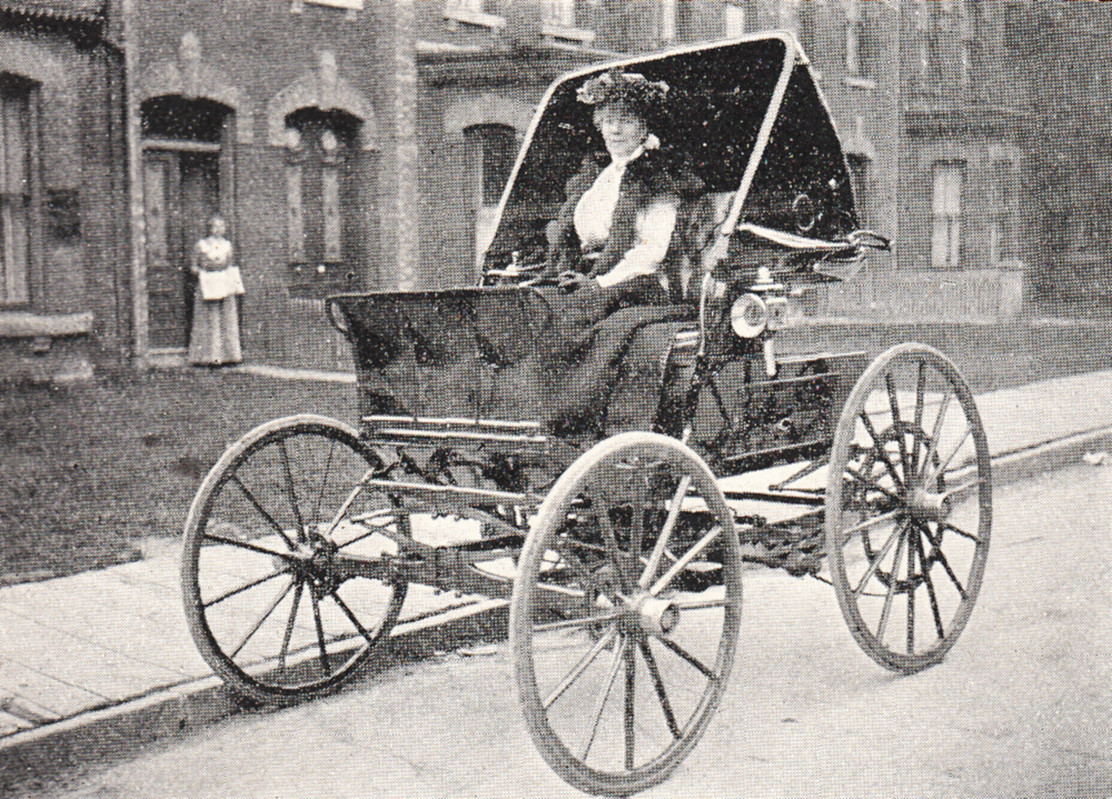 Black and white photograph of a well-dressed woman sitting in an open-topped electric car resembling a carriage.