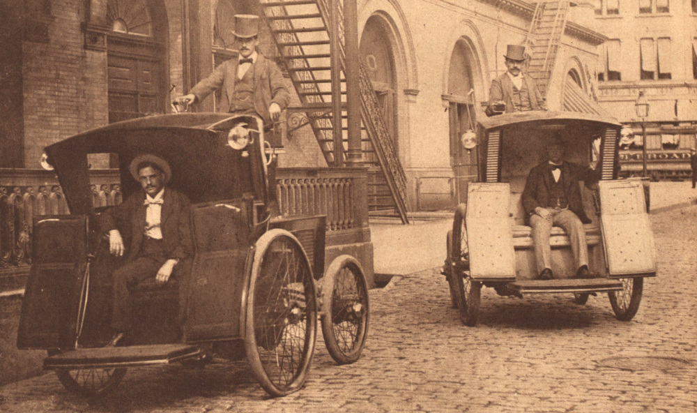 Two open-fronted electric taxicabs parked next to each other in a street scene. Each carries a passenger and a uniformed operator in a top-hat.