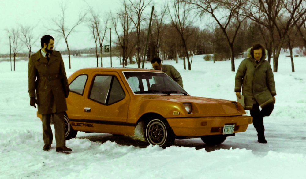Colour photograph of three men in coats gathered around an electric car in snowy countryside.