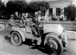 Black and white photograph of four men and a baby posed in an open-topped car, with six women and girls watching from behind a white picket fence in the background.