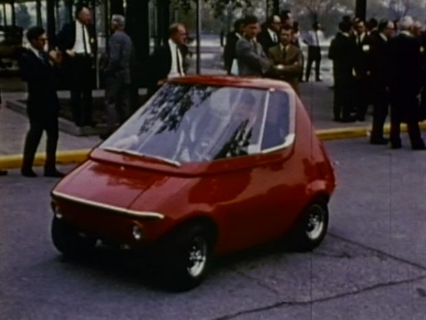 A small, bulbous vehicle parked in front of a crowd of men in business suits.