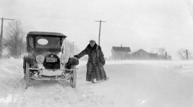 Black and white photograph of a man in a fur coat leaning against an automobile in deep snow. Farm buildings and fields are visible in the background.