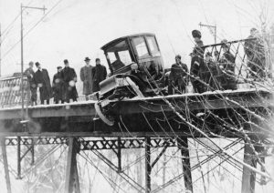 Black and white photograph of an electric car that has partially driven off a bridge in snowy weather. The car is surrounded by a crowd of police officers and onlookers.
