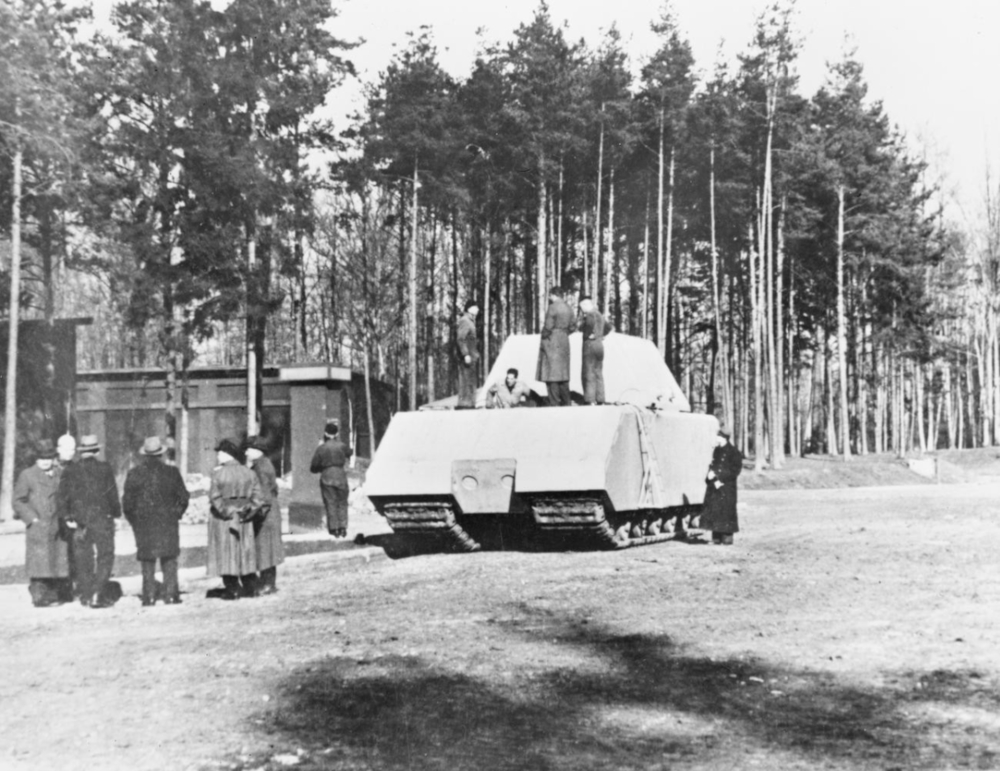 Black and white photograph of 12 men, some in military uniforms, standing on and around a very large angular armoured vehicle in a forest.