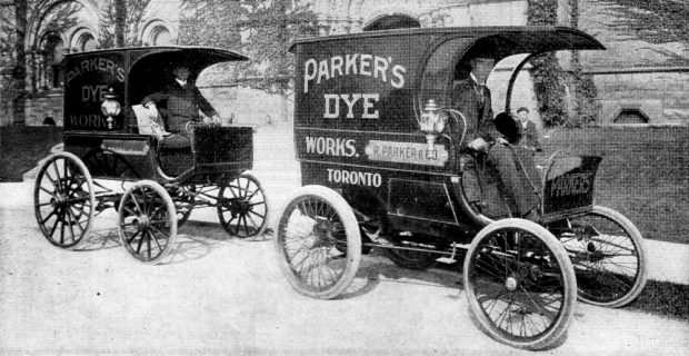 Two boxy, carriage-like motor vehicles are parked in front of a stone building, with drivers relaxing in their seats. Signs on both vehicles read PARKER'S DYE WORKS.