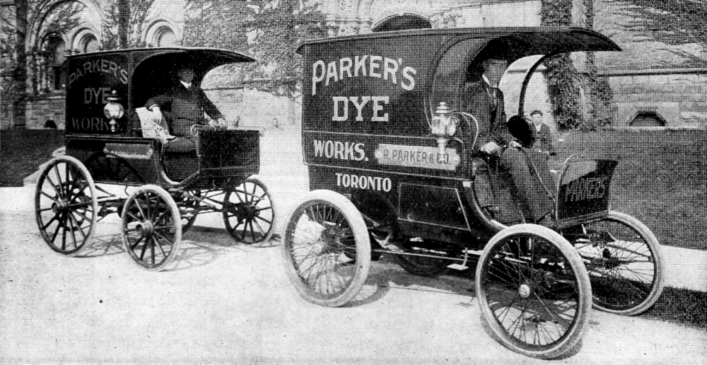 Two boxy, carriage-like motor vehicles are parked in front of a stone building, with drivers relaxing in their seats. Signs on both vehicles read "PARKER'S DYE WORKS".