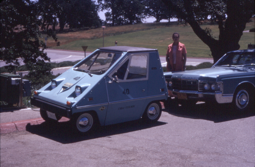A very small two-seater electric car is plugged in at a curbside charging station, with gardens in the background. A woman is standing behind the car, looking at the camera.