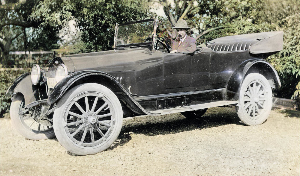 Colourized photograph of a man in a suit sitting in the front seat of a 1920s-style convertible automobile.