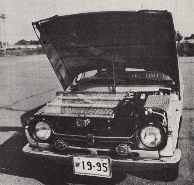 Black and white photograph of a car with a Japanese license plate with its hood open. Its engine compartment is packed full of hundreds of rectangular battery cells.