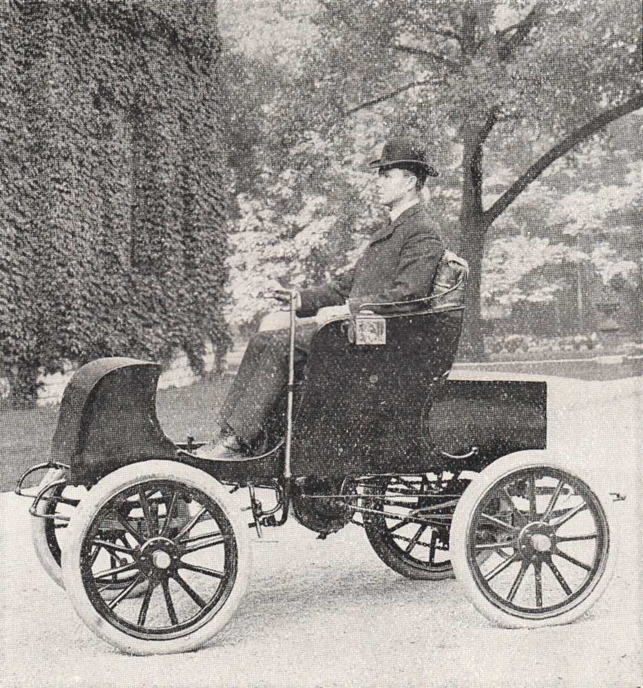A well-dressed man seated in an open-topped two-seater electric car in front of a stone building covered in ivy.