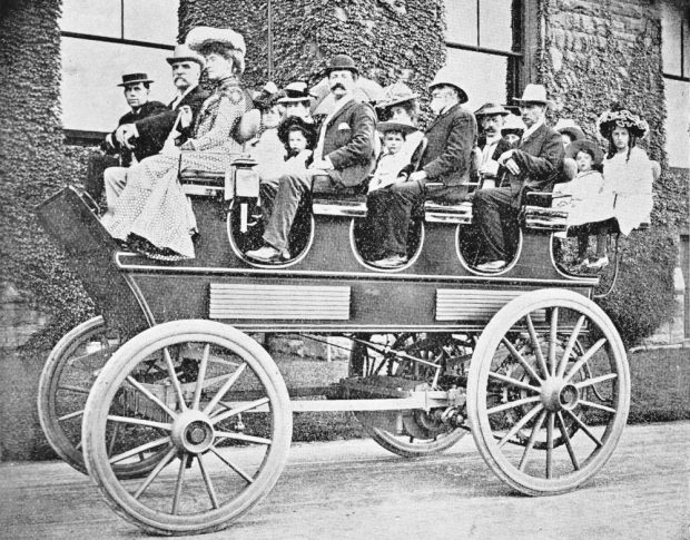 Seventeen people of all ages in Edwardian dress sit in an open-topped electric carriage.