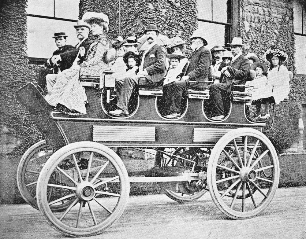 Seventeen people of all ages in Edwardian dress sit in an open-topped electric carriage.