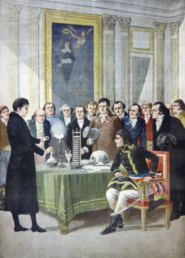 Colour woodcut of a man demonstrating sparking electrical apparatus to a group of men in 19th-century formal dress, including a seated Napoleon Bonaparte.